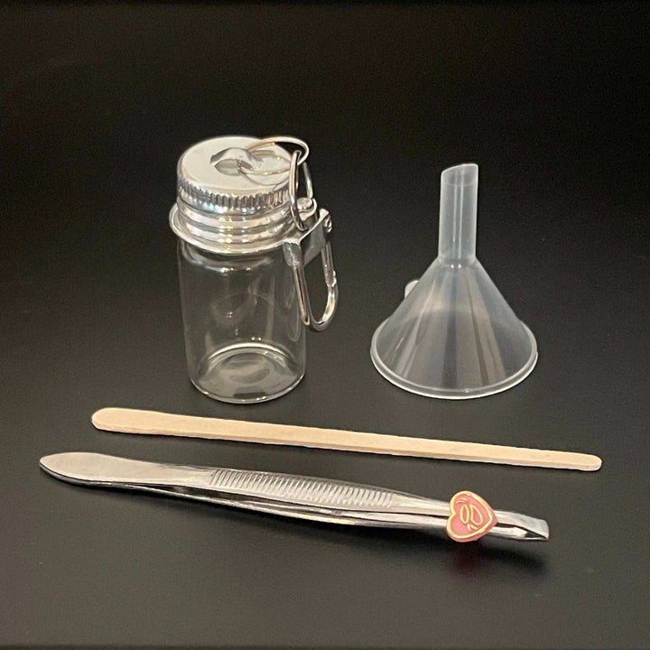 A small clear glass jar for dog or cat fur or ashes with a silver metal clasp lid, shown with a clear plastic funnel, a wooden spatula, and a metal tool on a dark background.