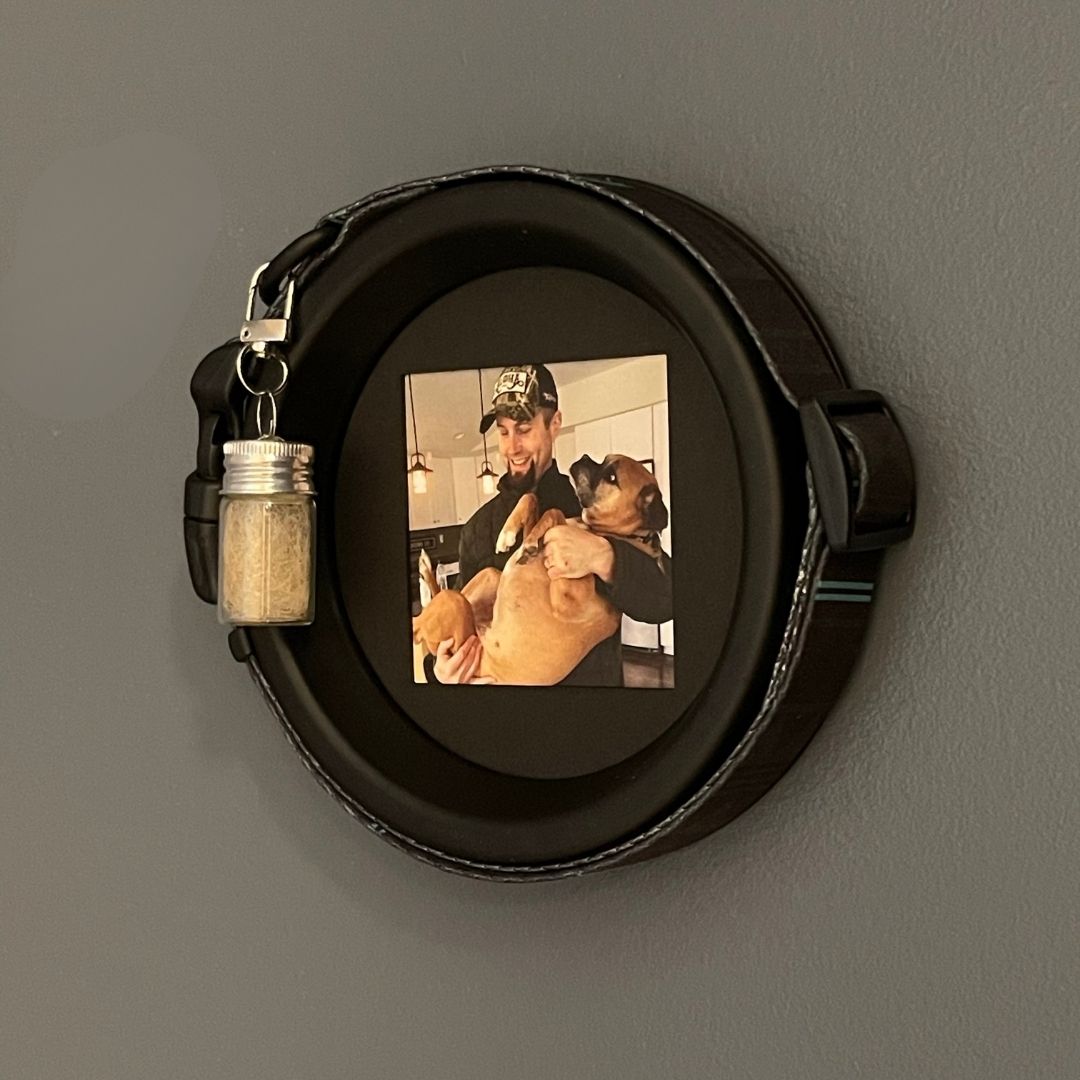 A black circular pet collar display frame with a small hanging glass jar charm filled with dog fur, displayed on a gray wall. 